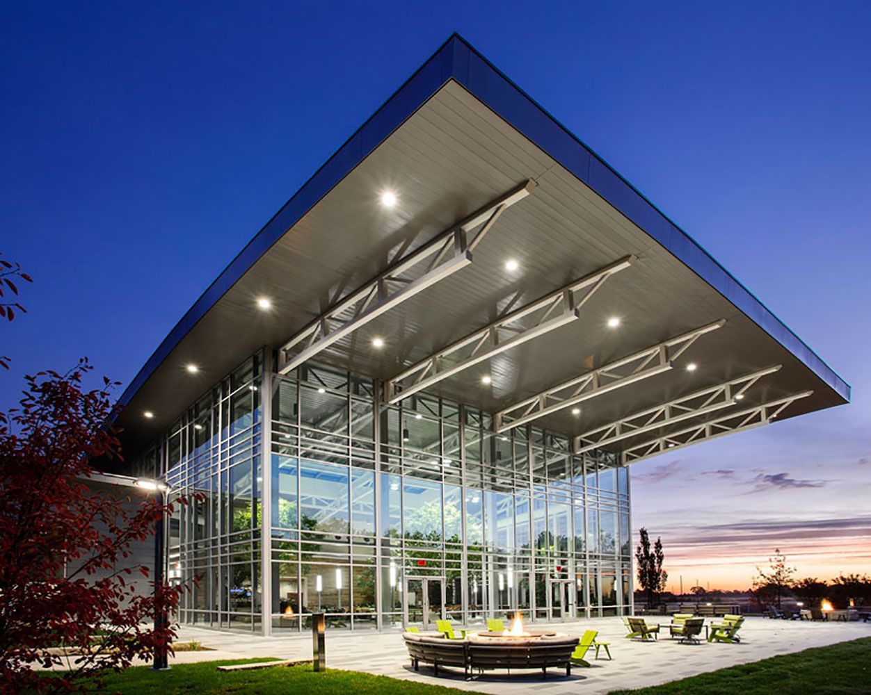 Modern glass pavilion with a dramatic angular roof at dusk, lit from within and surrounded by outdoor seating.
