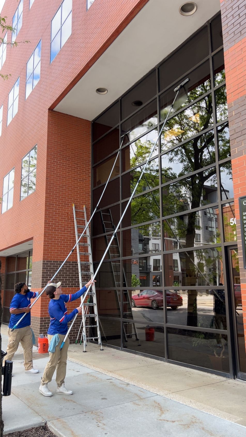 Workers on a sidewalk using ladders to clean a tall glass building entrance