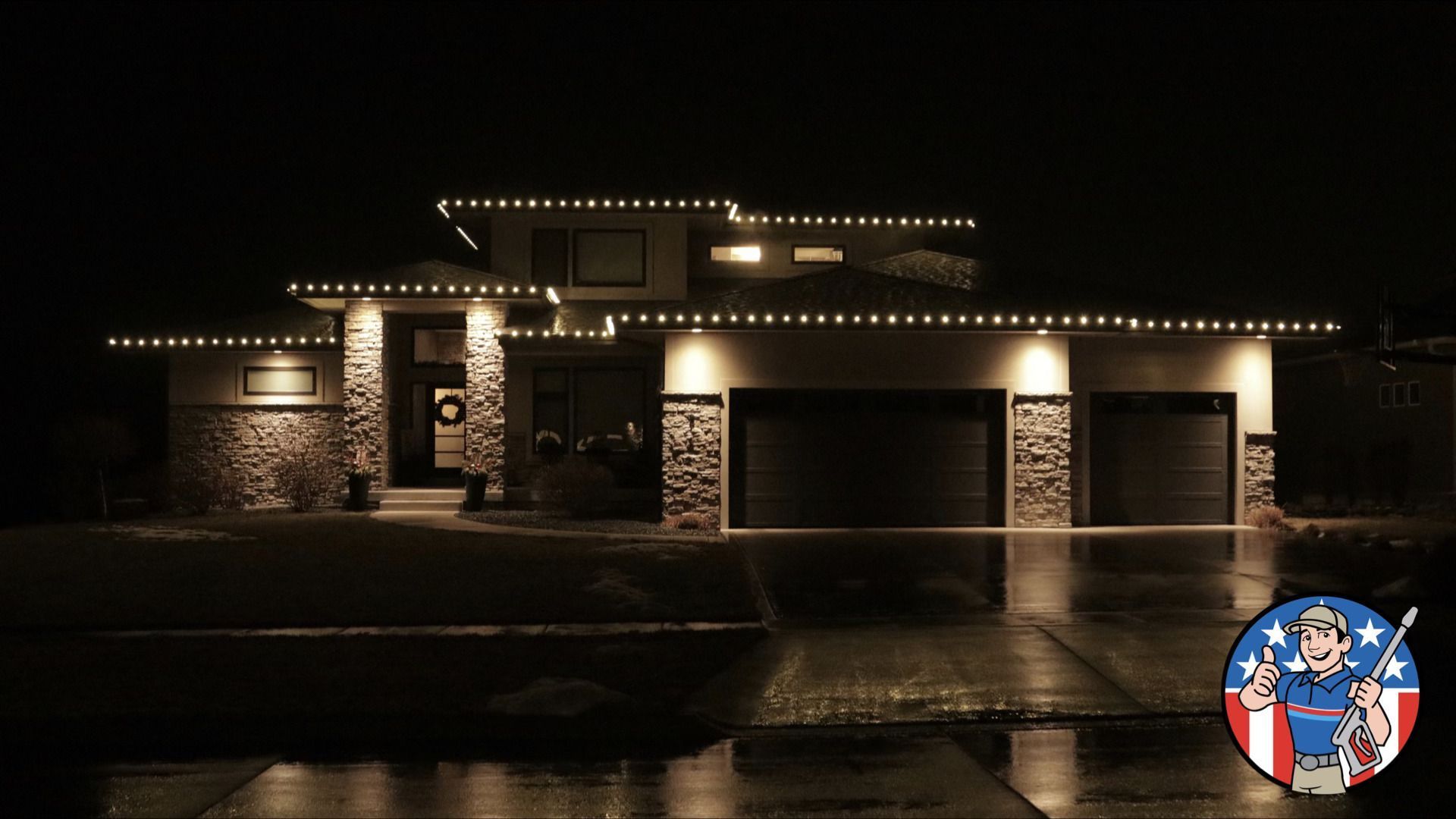 Nighttime view of a lit suburban house with warm roofline lights, stone facade, and wet driveway