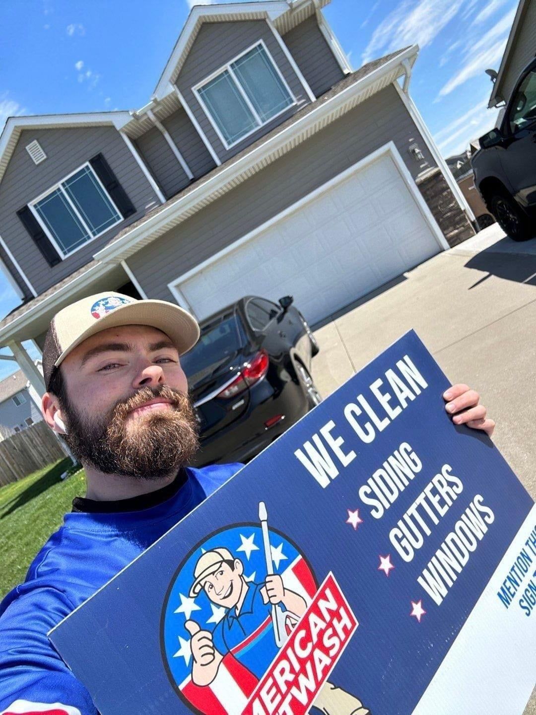 Man in blue shirt holding a sign for pressure washing near a house and parked cars