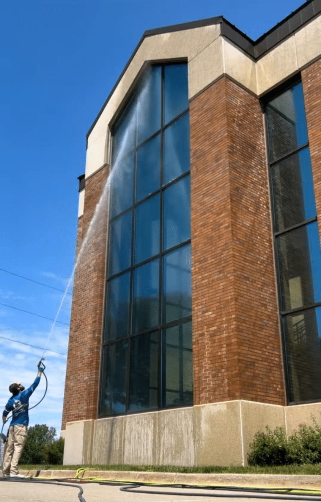 Tall brick building with blue-tinted glass windows and a worker using a hose at the lower left