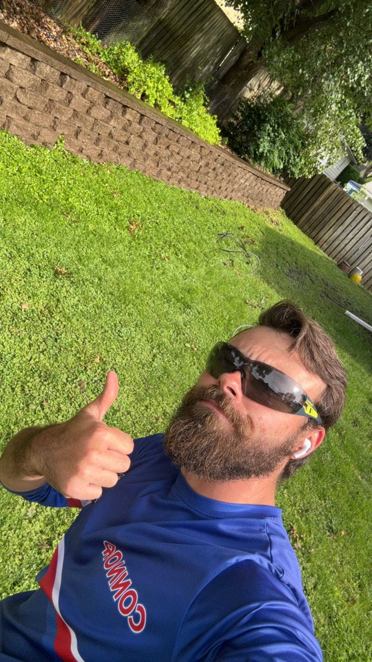 Man giving a thumbs-up selfie on a grassy lawn, wearing sunglasses and a blue shirt.