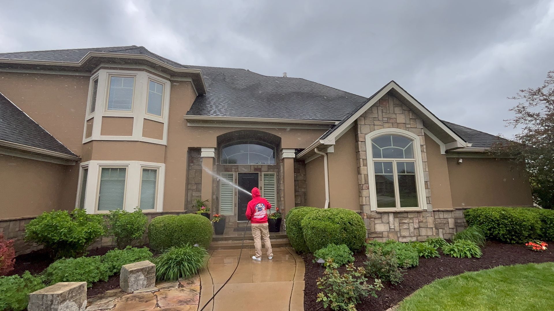 A large beige suburban house with a person in a red jacket standing on the front walkway.