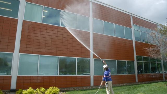 Person spraying water on a brick office building with large windows and green shrubs below.