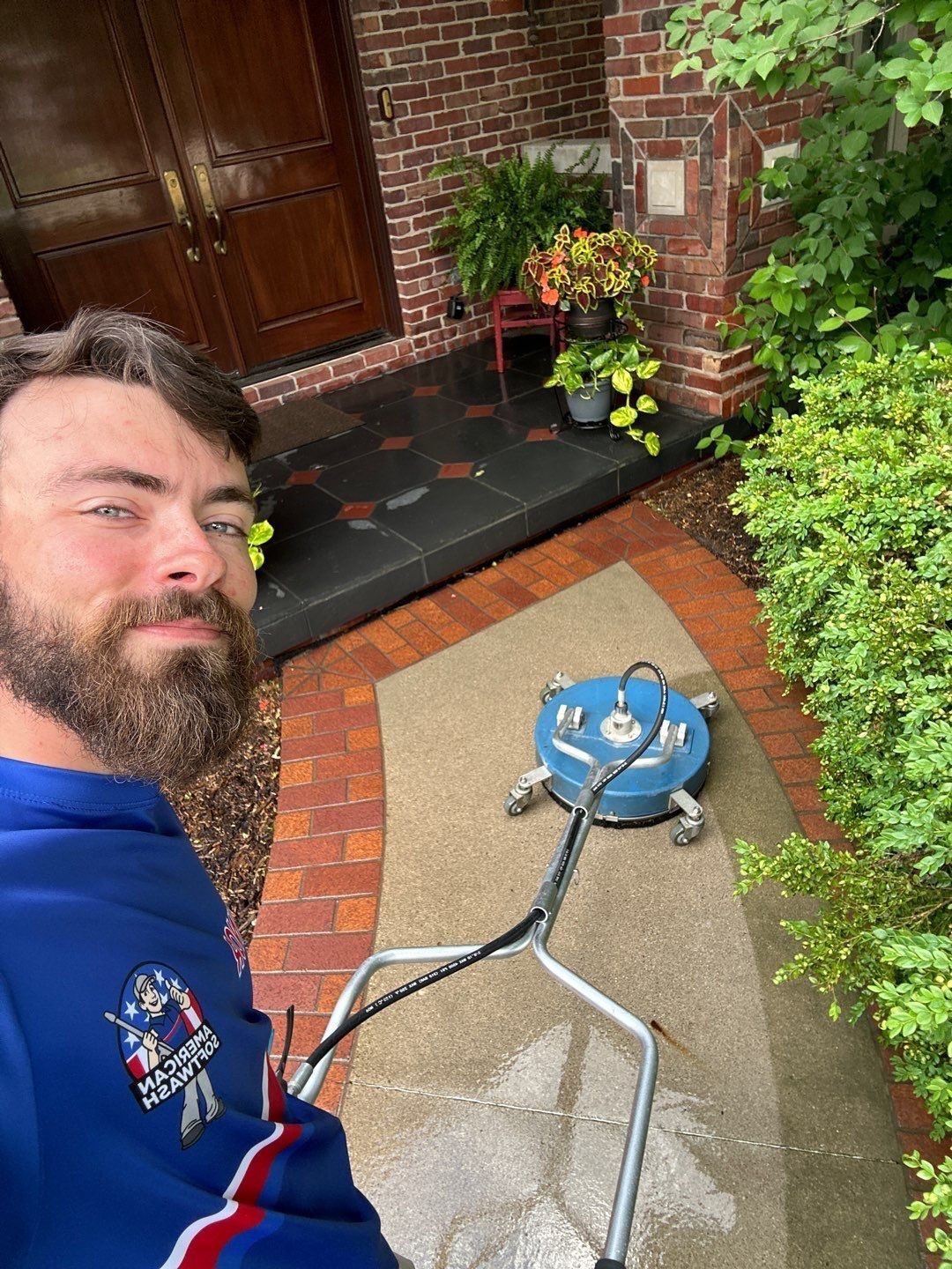 Person smiling beside a pressure washer on a brick patio by a front door and garden greenery