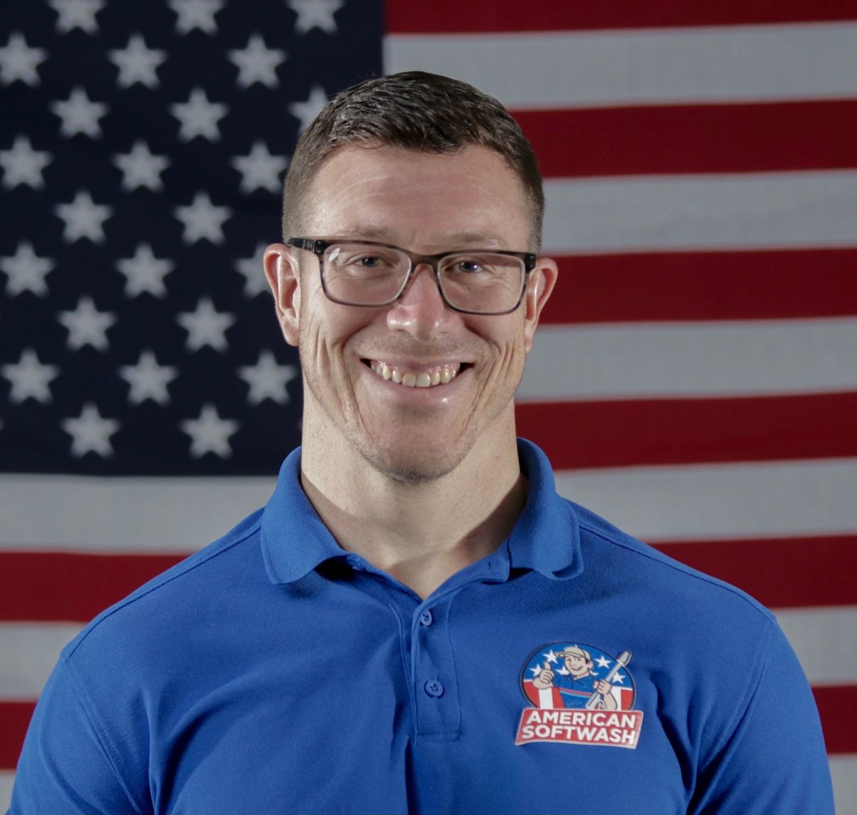 Smiling man in blue polo with American Dream Farms logo, standing before a U.S. flag backdrop