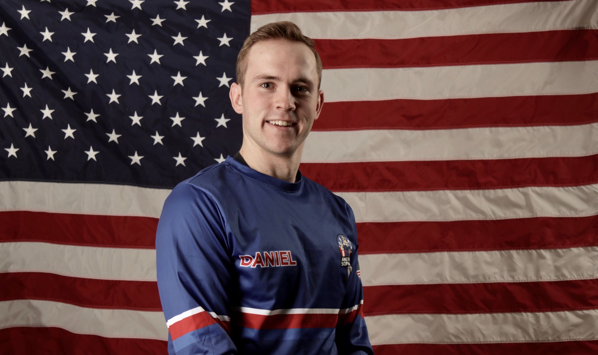 Man in a blue sports jersey standing before a large American flag backdrop
