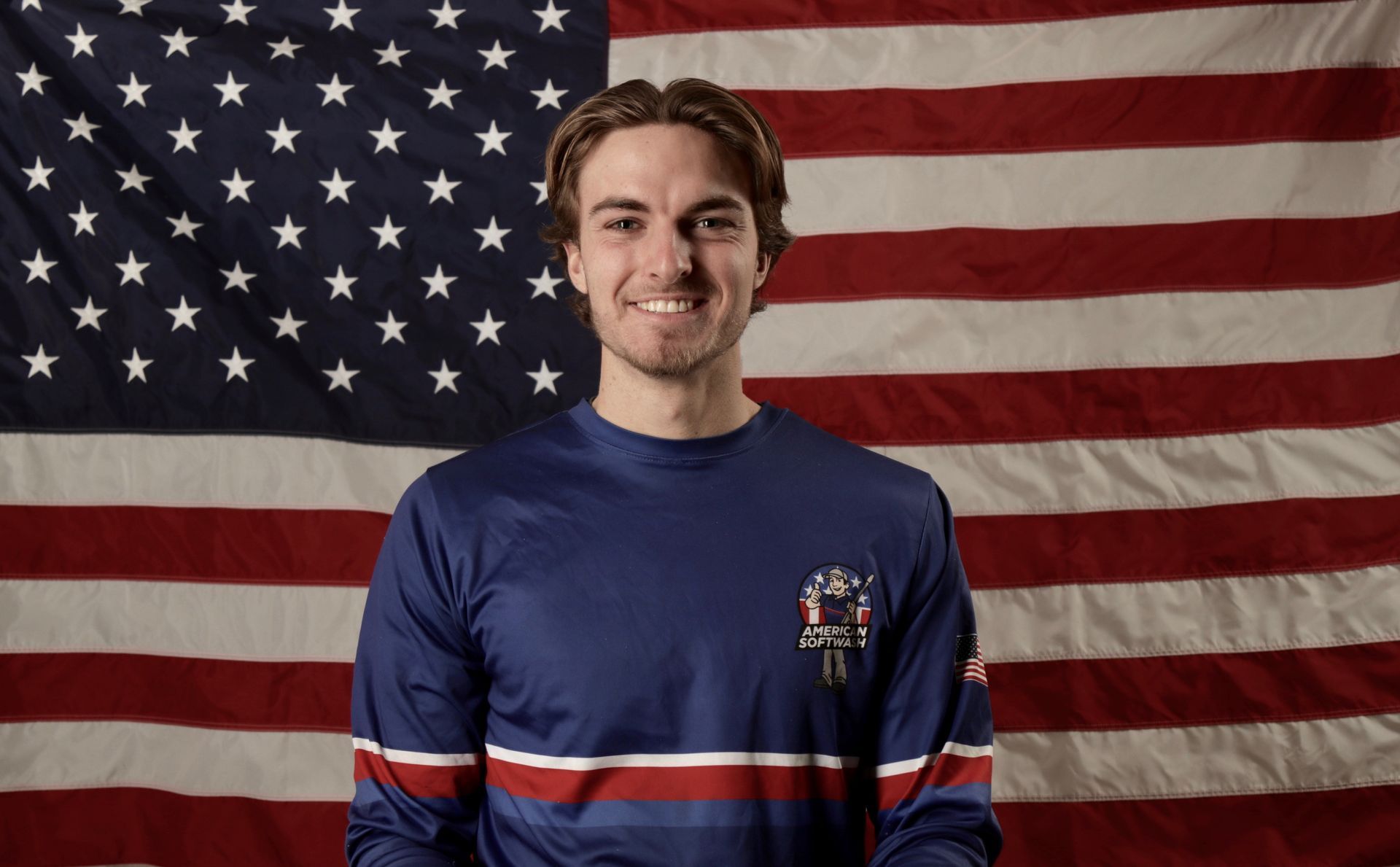 Smiling person in a blue shirt standing before a U.S. flag backdrop