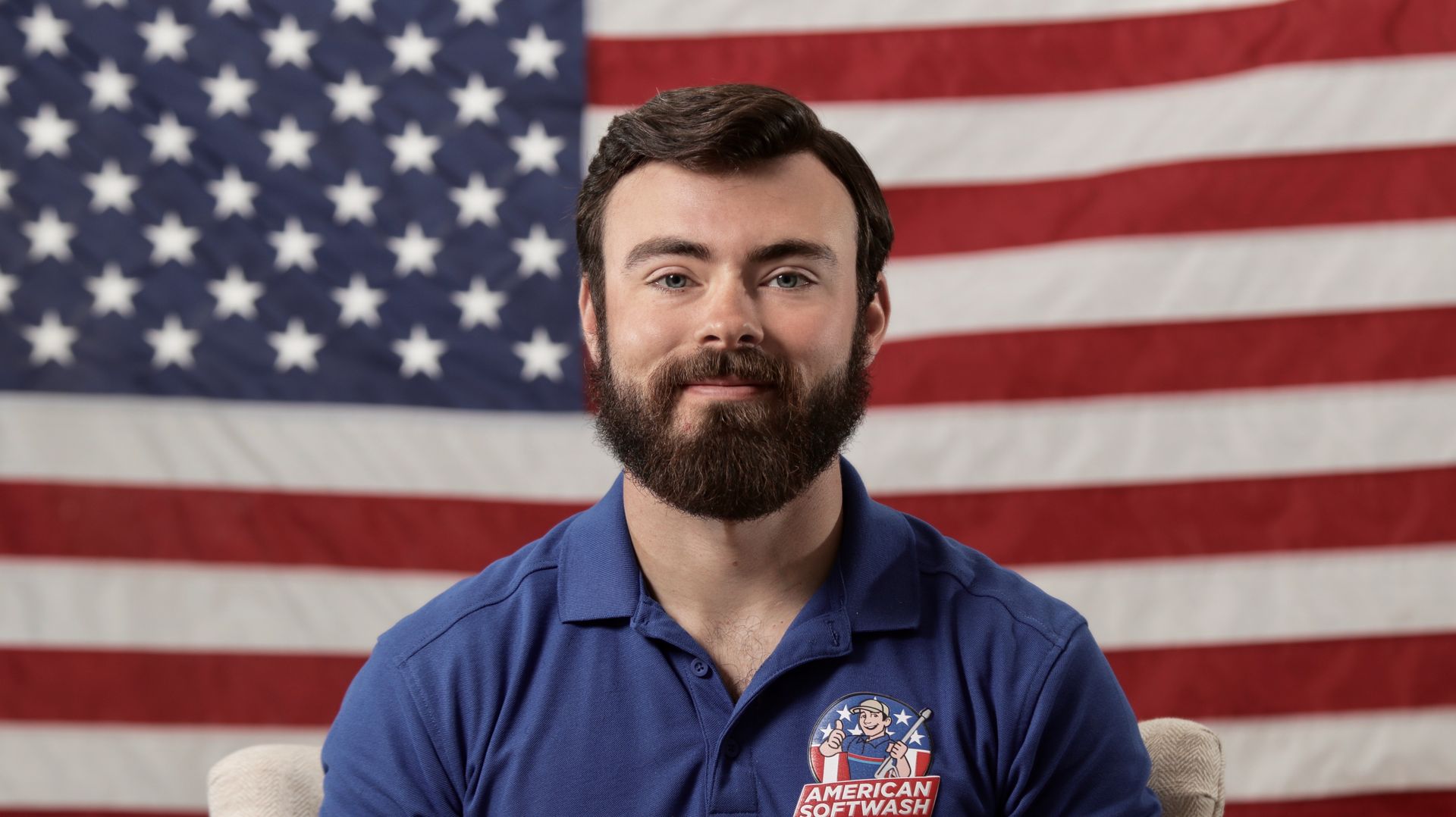 Portrait of a bearded man in a blue polo, seated before a large American flag backdrop