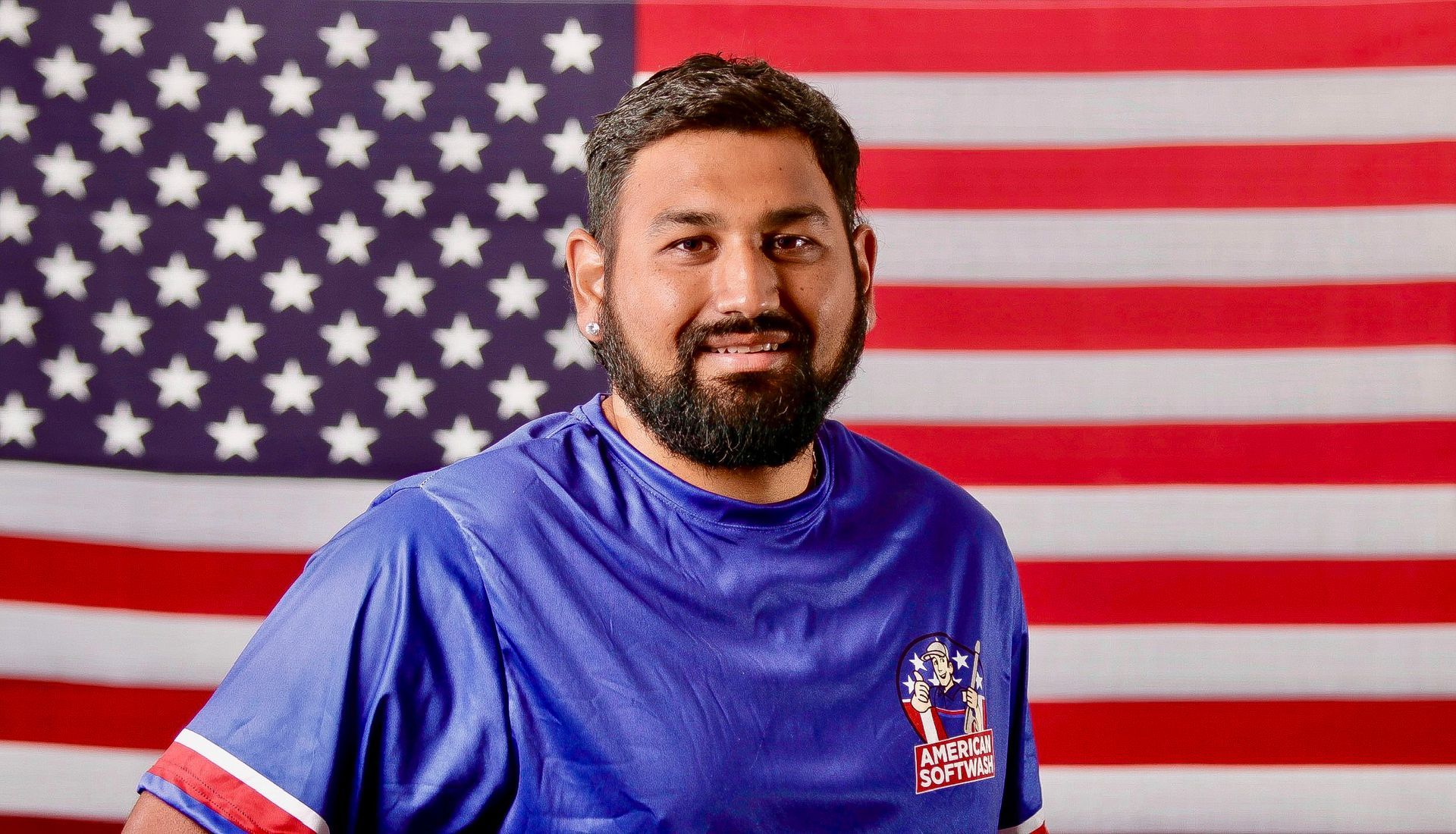 Man in a blue shirt standing before a large American flag, smiling at the camera.