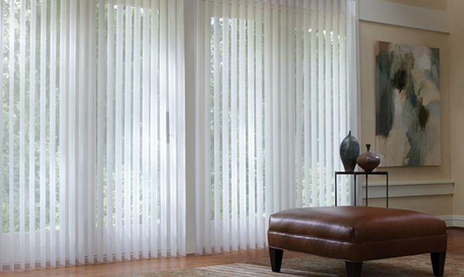 A living room with white vertical blinds and a brown ottoman.
