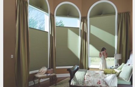 A woman is standing in a bedroom with arched windows and honeycomb blinds.