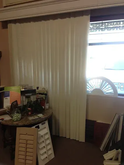 A room with white blinds and a fan in front of a window.