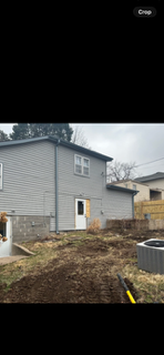 Backyard beside a gray house with a white door, bare yard, and neighboring homes under a cloudy sky