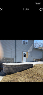 Side view of a house with a stone retaining wall, white siding, and a small porch steps under clear sky