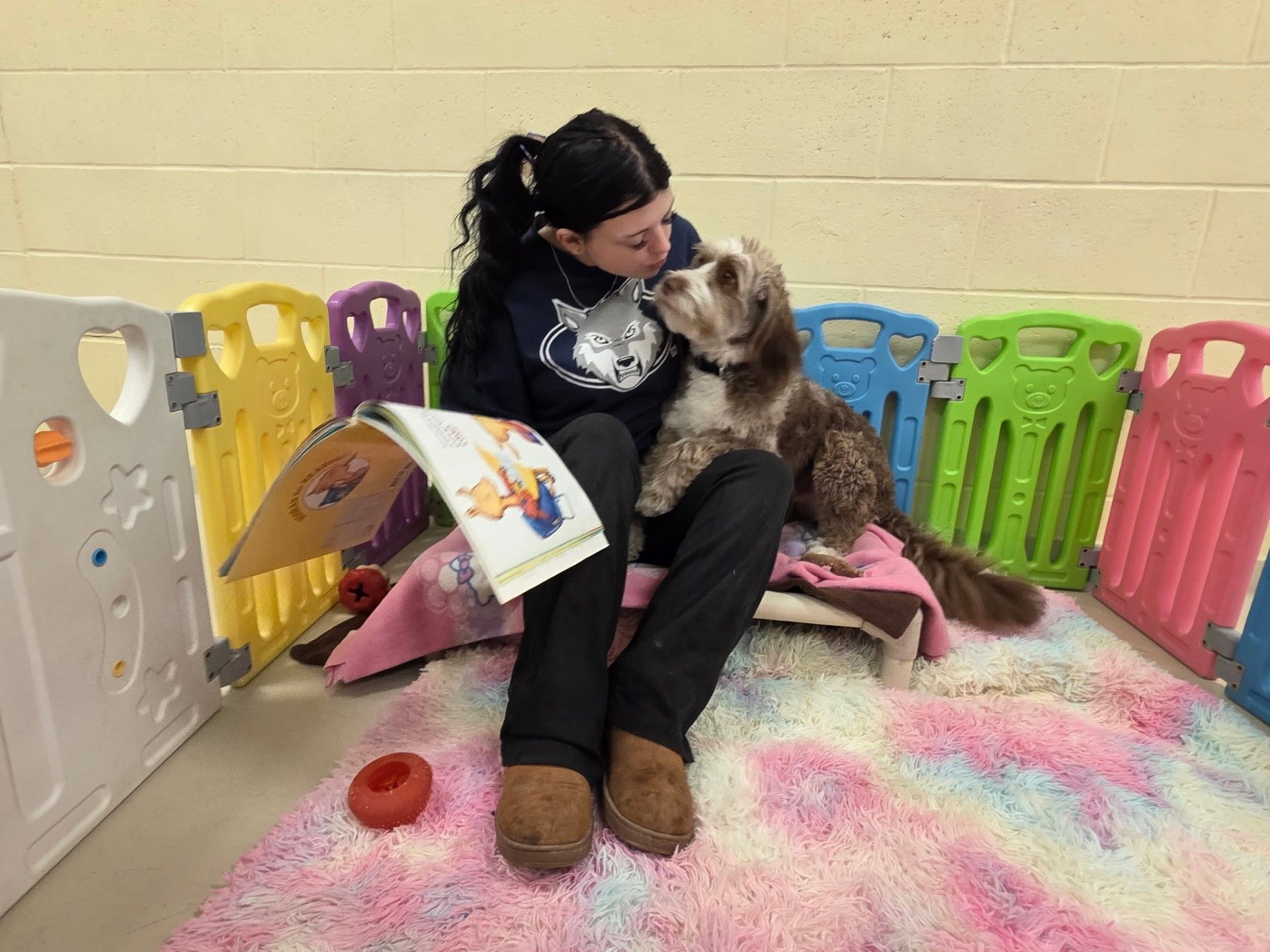 Woman cuddles a dog while reading a book in a colorful playpen