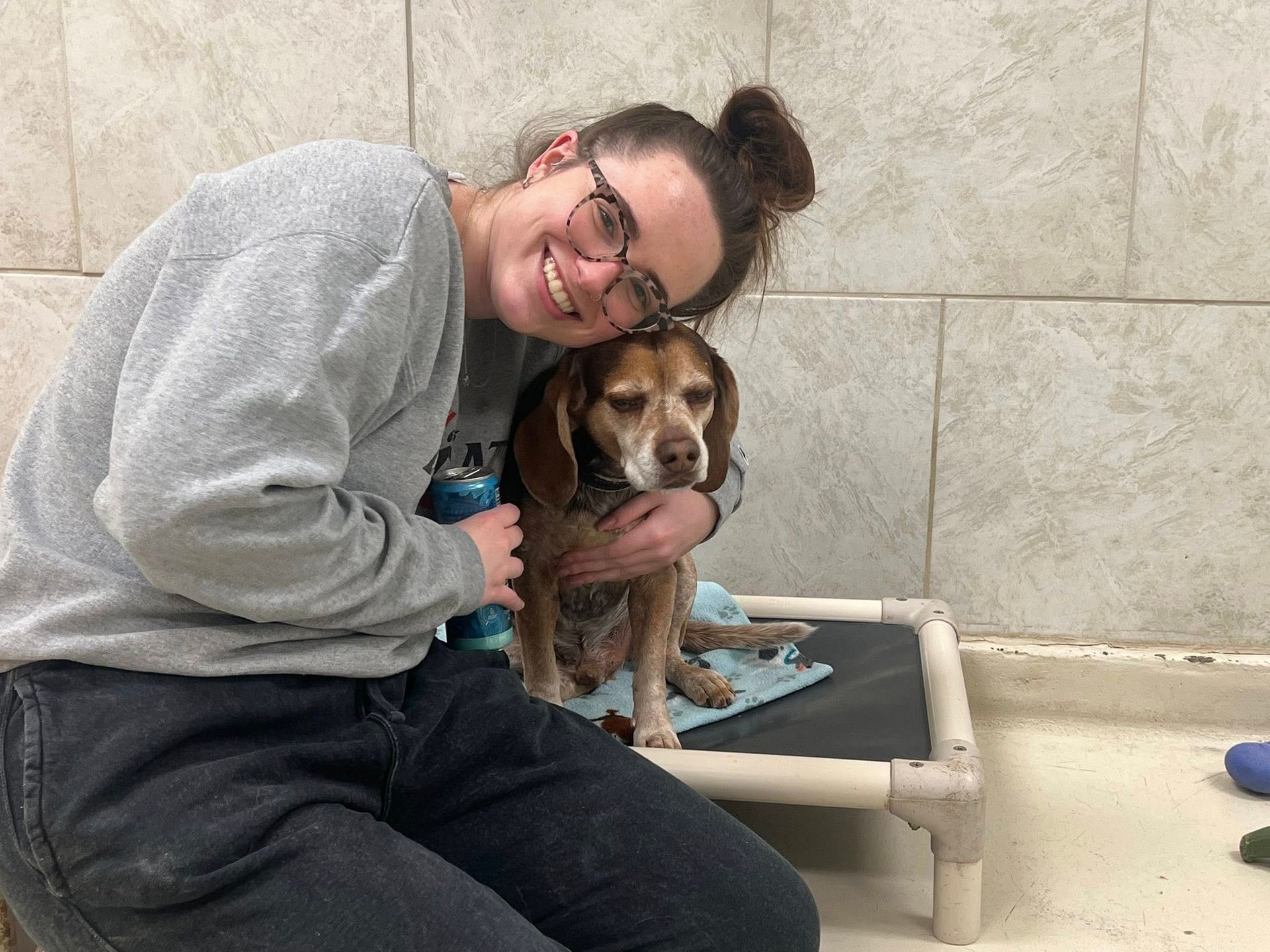 Woman in glasses smiles, hugging a beagle dog on a dog bed; indoor setting