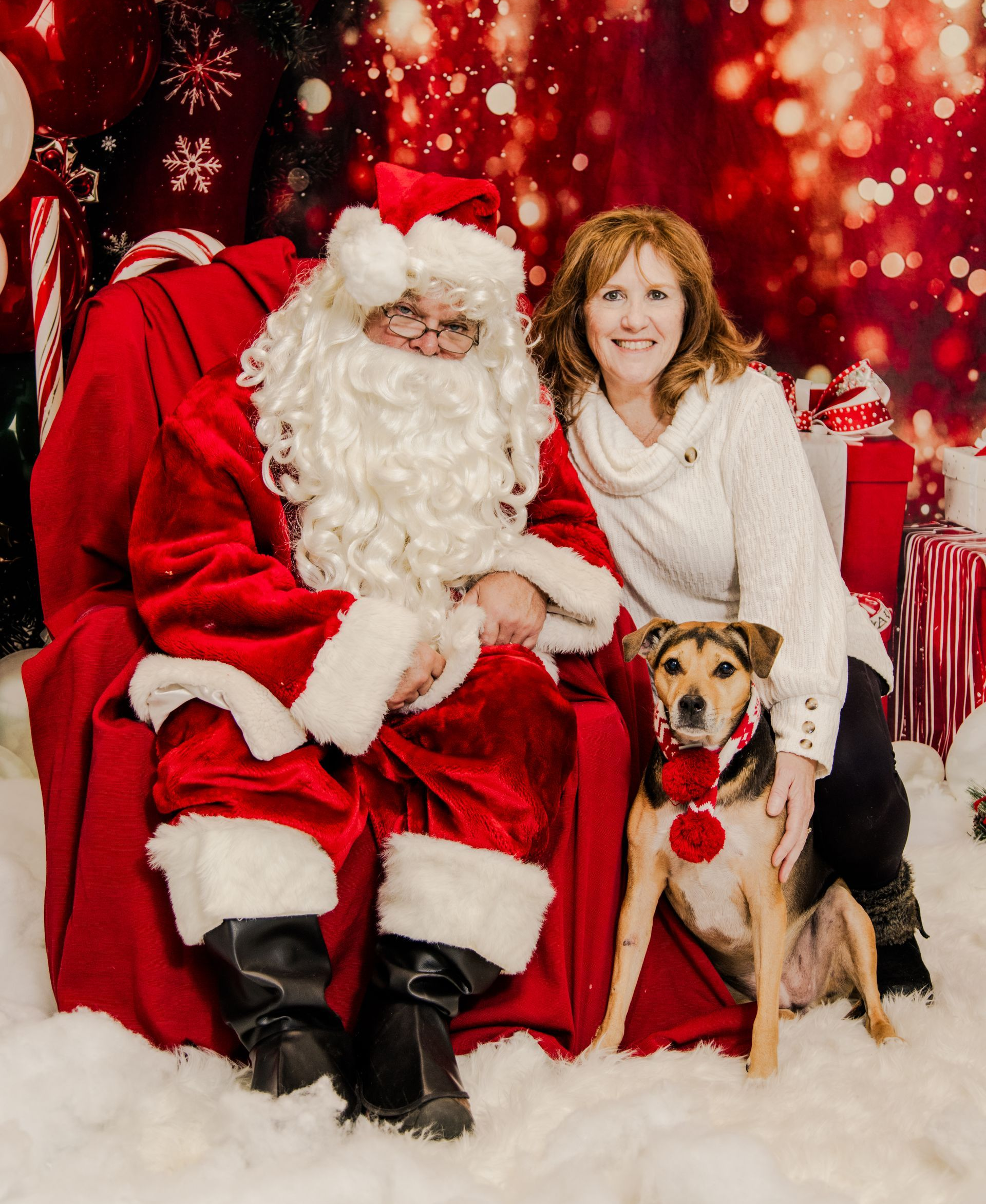 Santa Claus seated with a woman and dog; all posing for a Christmas photo. Red, white, and snowy background.
