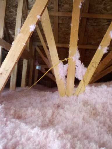 Attic framing with pink insulation on the floor and rafters, showing wooden beams and yellow wiring.