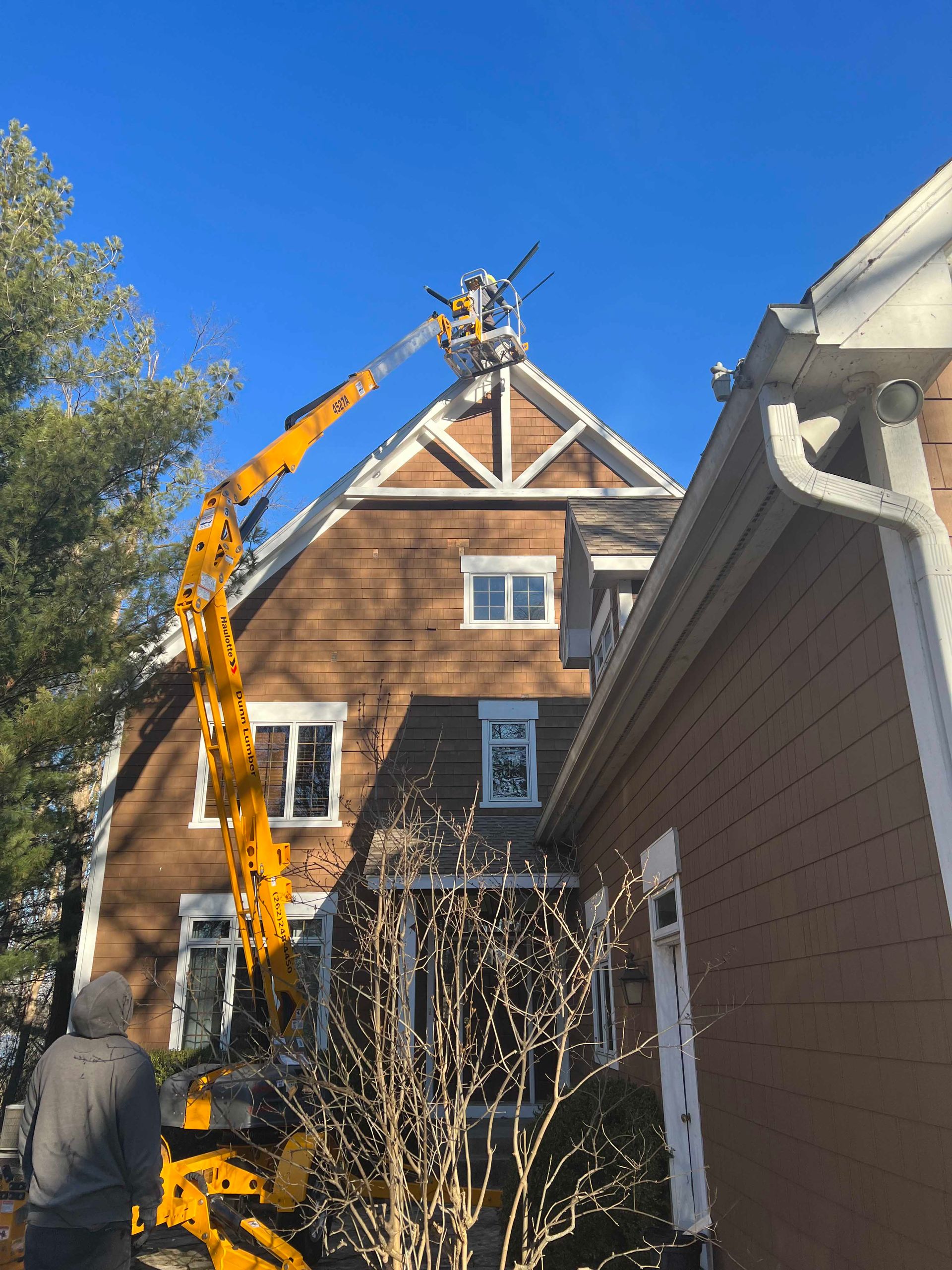 Yellow crane lifts roof beams on a house under construction beside a brown shingled home.