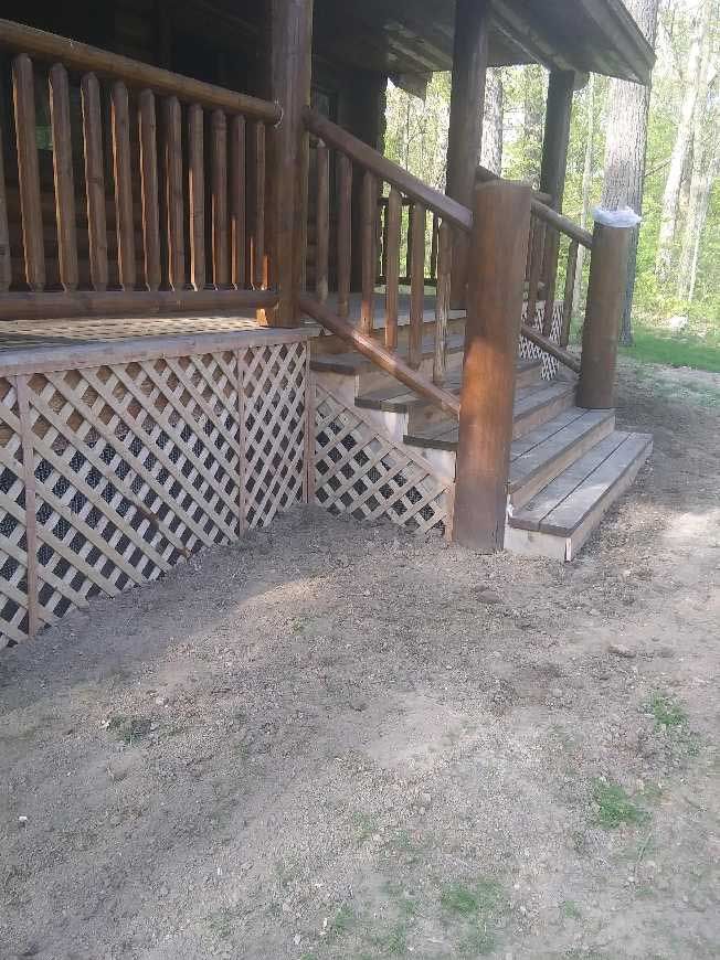 Log cabin porch with wooden stairs, railing, and lattice skirting on a dirt yard