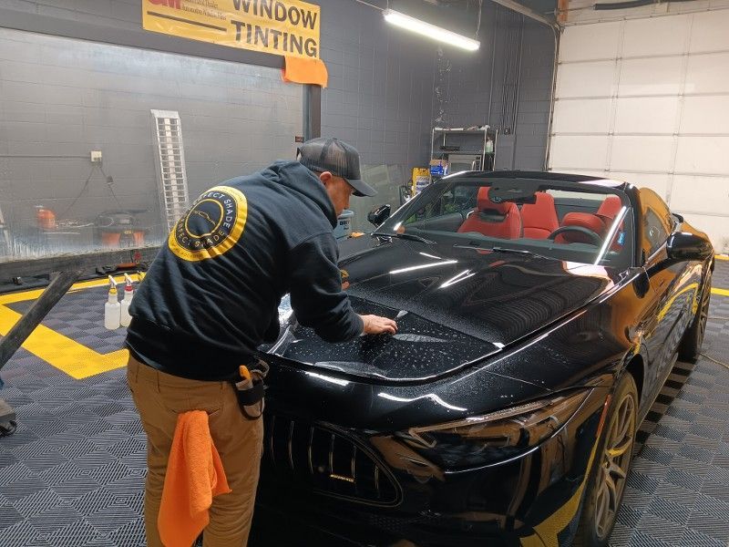 A man is applying window tinting to a black car in a garage.