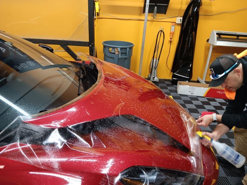 A man is applying a protective film to the hood of a red car.
