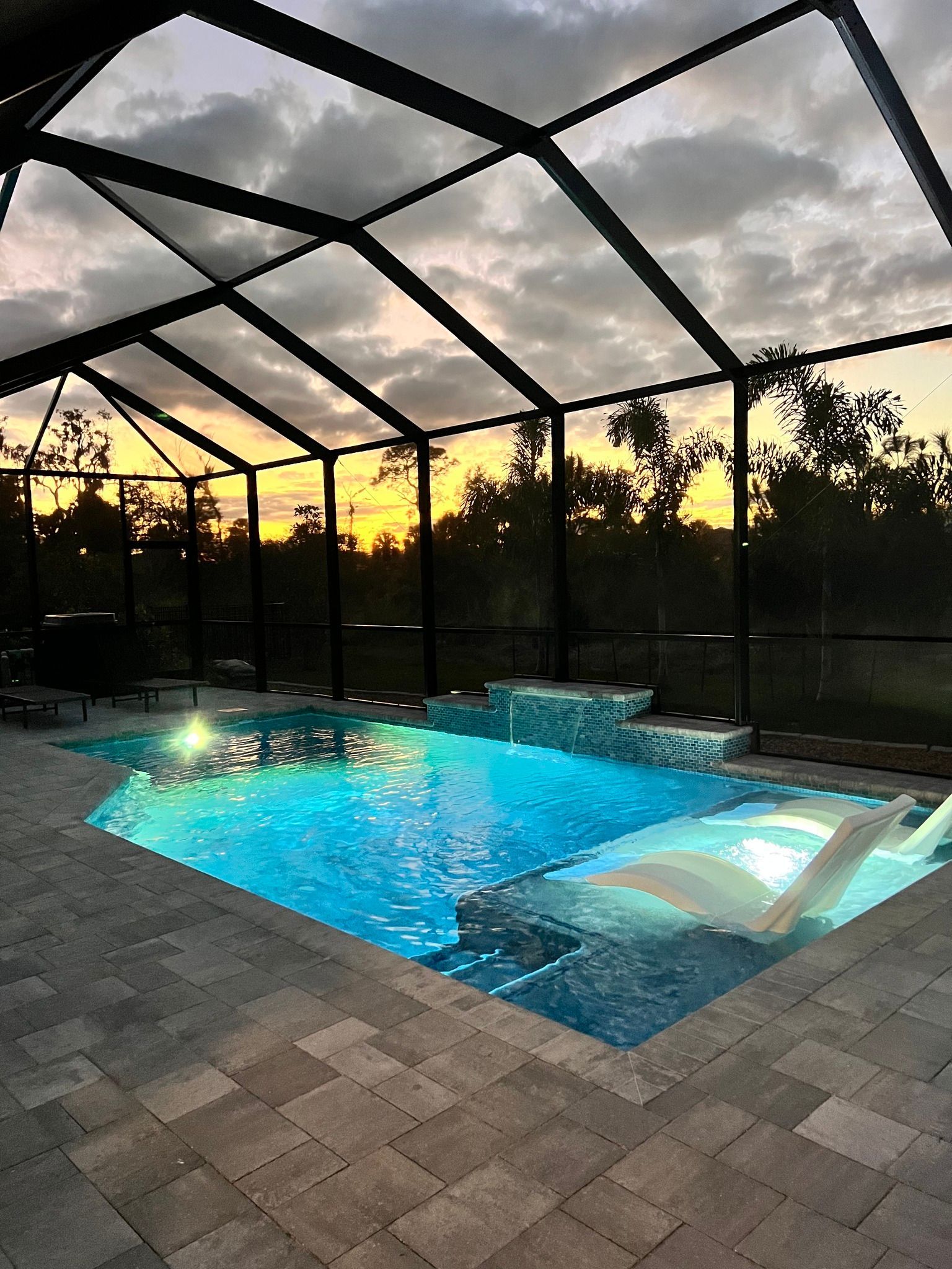 A large swimming pool is surrounded by a screened in area at sunset