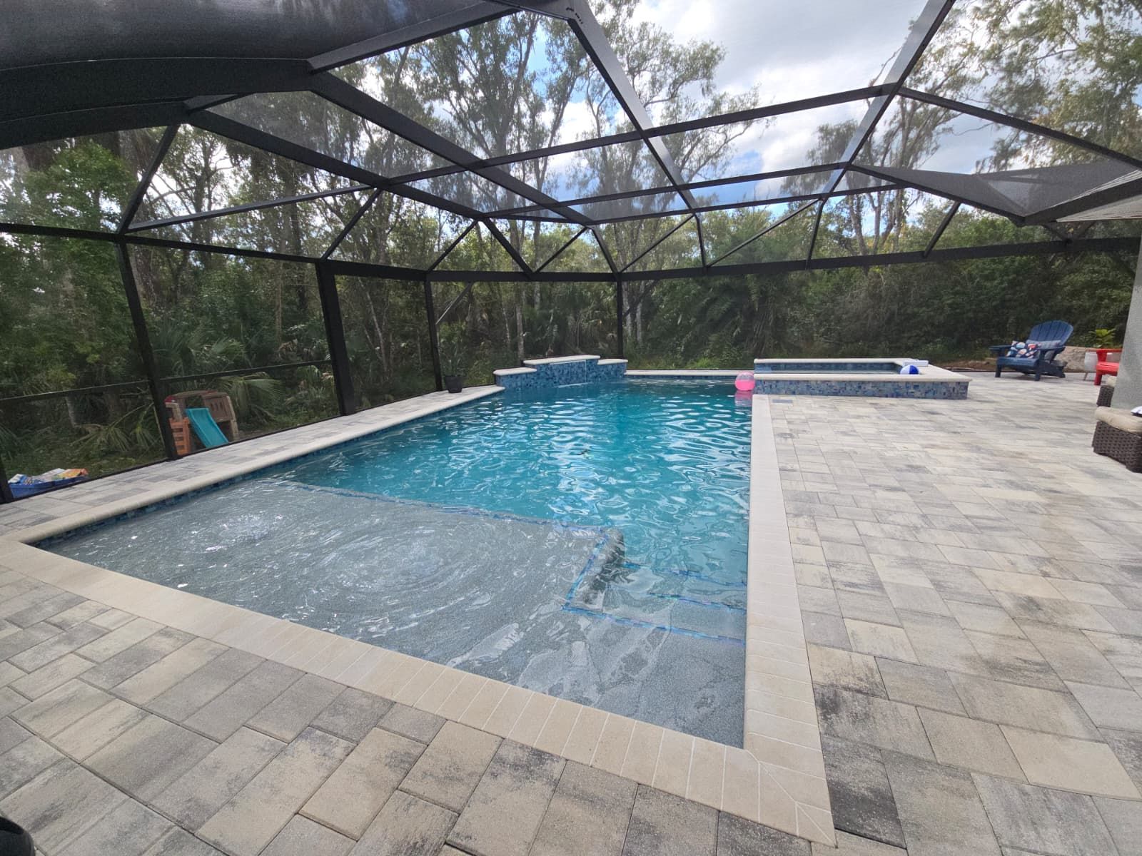 A sunlit, rectangular swimming pool with a Baja shelf, blue water, and a screened enclosure on a stone patio.