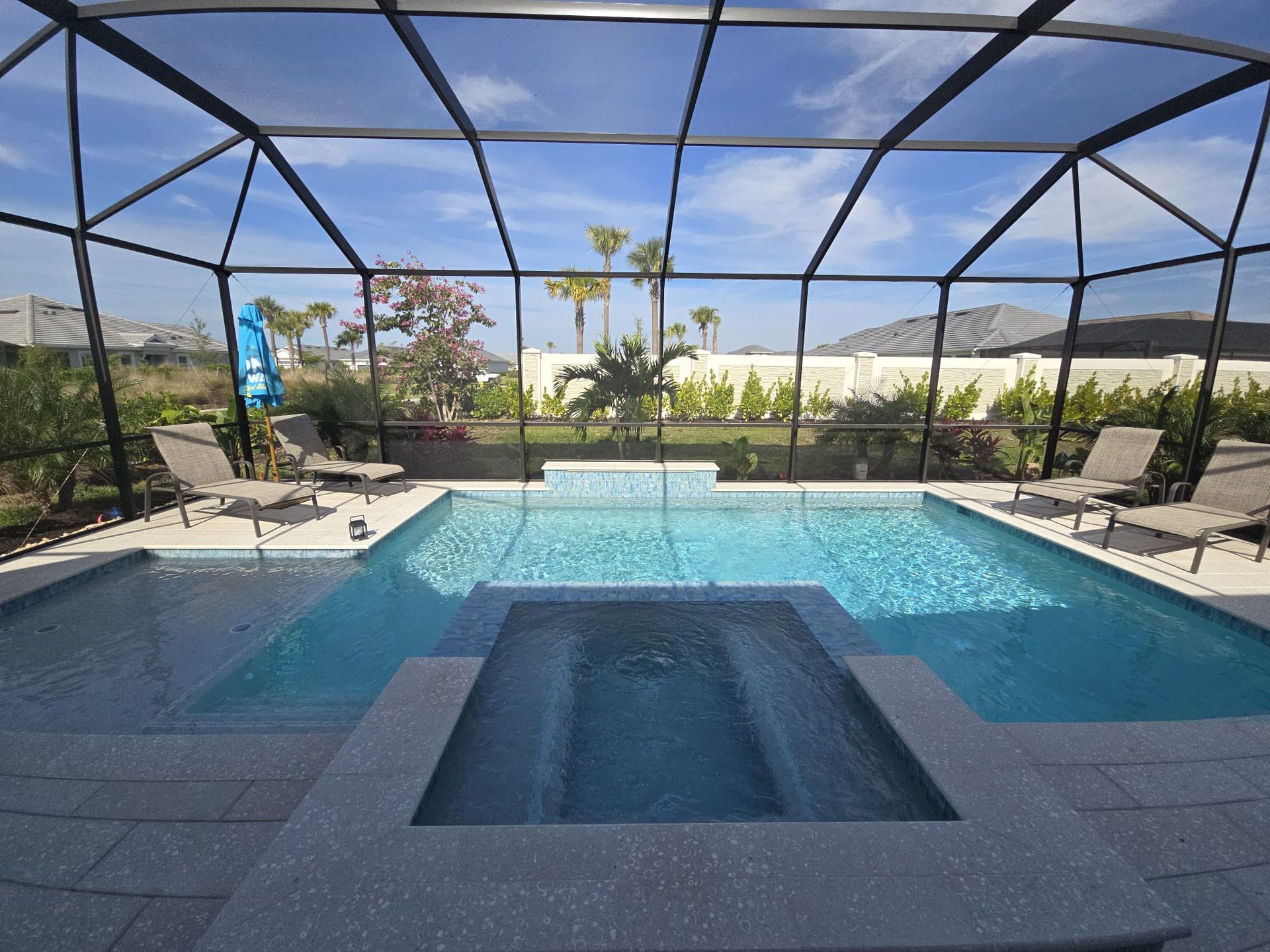 A sunny backyard swimming pool and spa enclosed by a dark mesh screen, with lounge chairs on a stone patio.