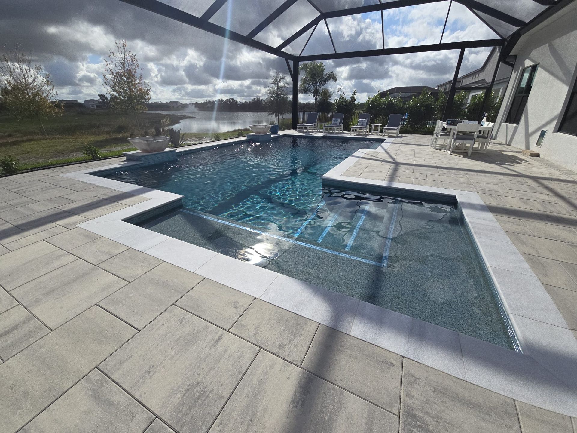 A backyard swimming pool with a screened enclosure, pavers, and a view of a lake under a partly cloudy sky.