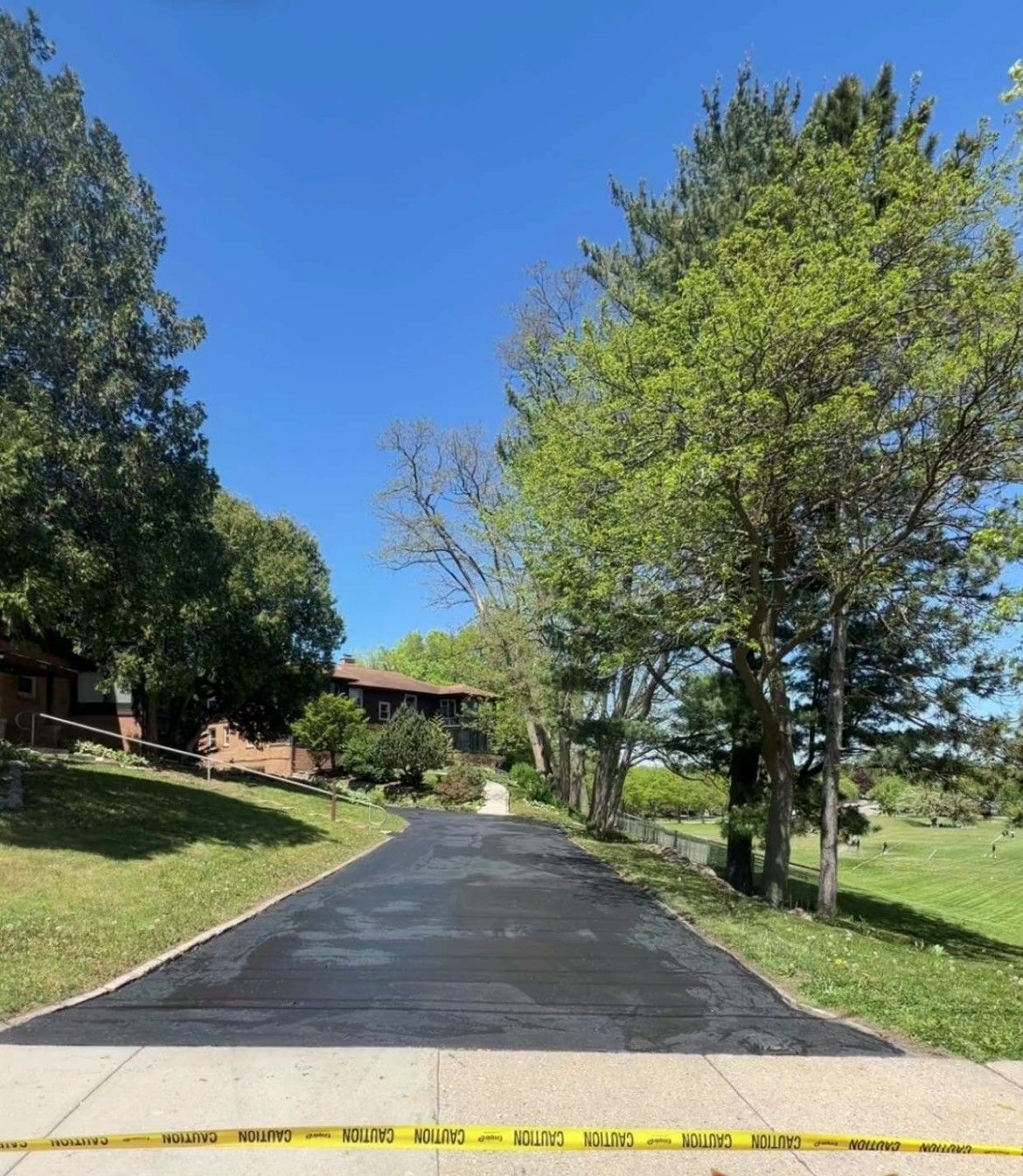 Paved driveway uphill to a house, lined with trees and grass, with yellow caution tape across the bottom.