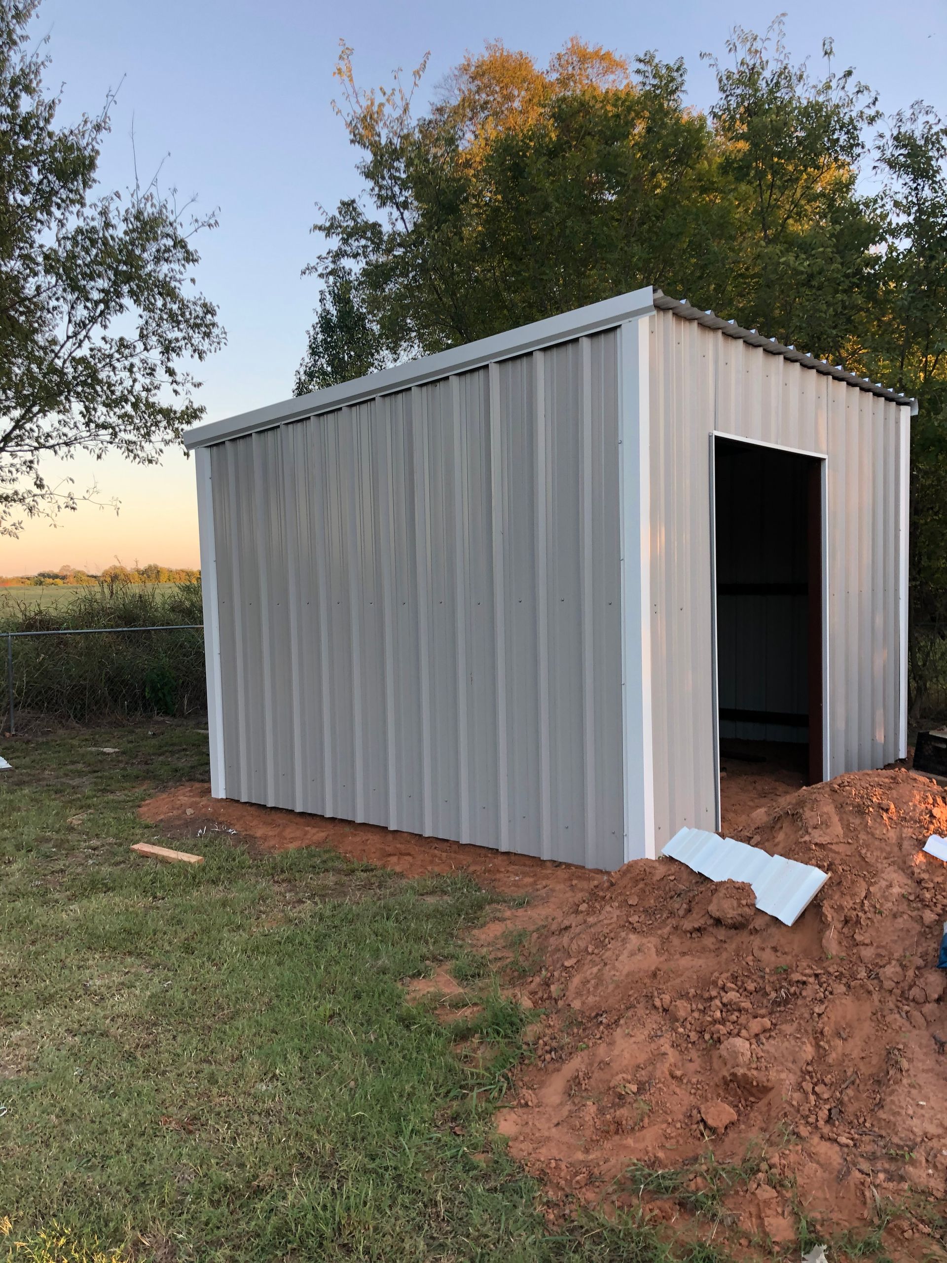 Small gray metal shed on bare dirt at sunset, with an open doorway and grassy field behind it