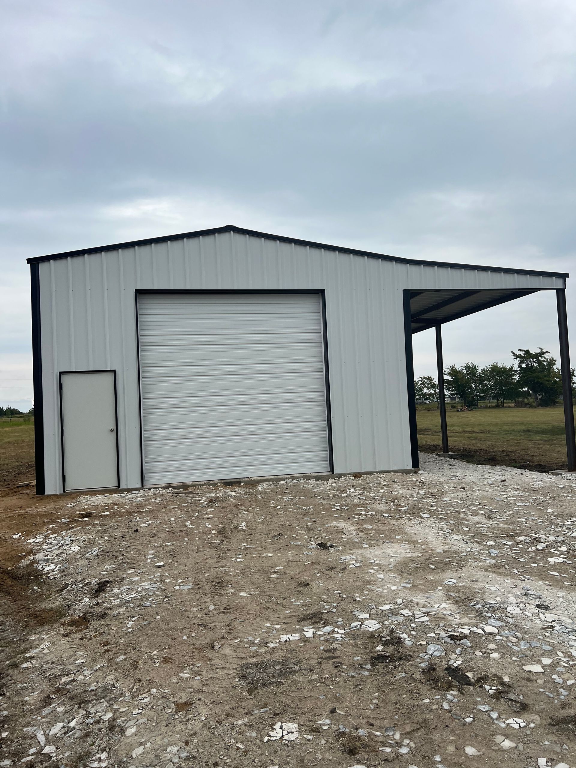 Small gray metal barn with a roll-up door and side lean-to on a gravel lot under cloudy skies