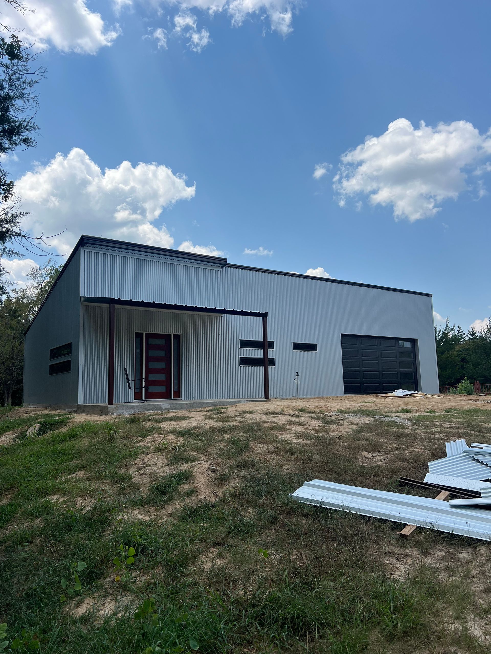 Metal-sided building on a grassy slope under a blue sky with clouds, with construction materials in the foreground.