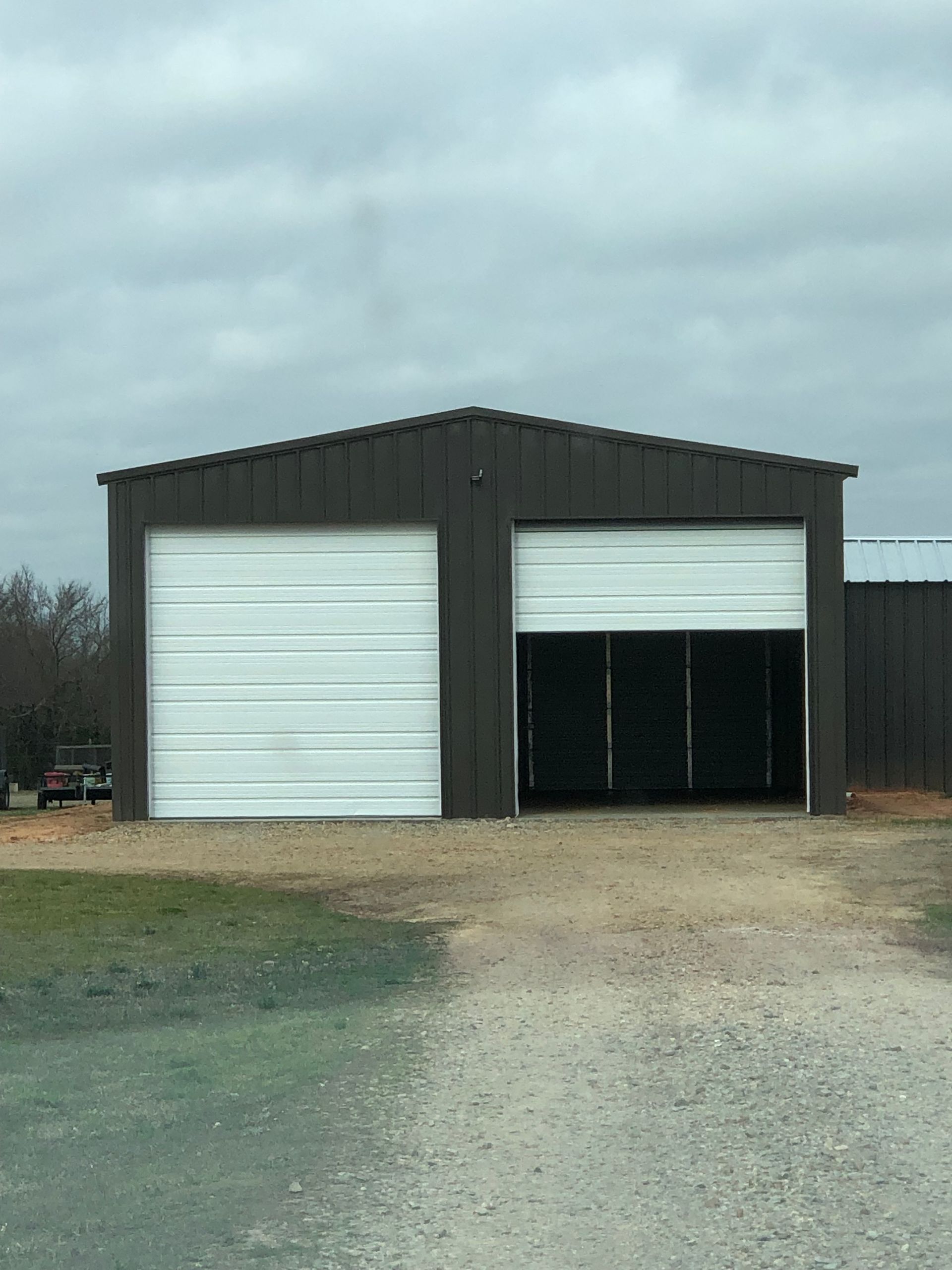 Dark metal garage with two large white doors, one open, on a gravel lot under a cloudy sky