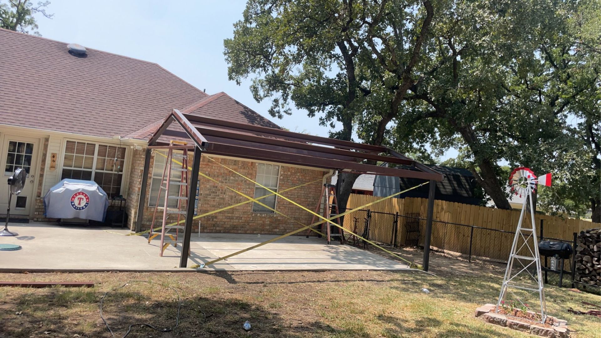 Backyard patio with a metal carport frame, stone house, and a ladder near a tree on the right.