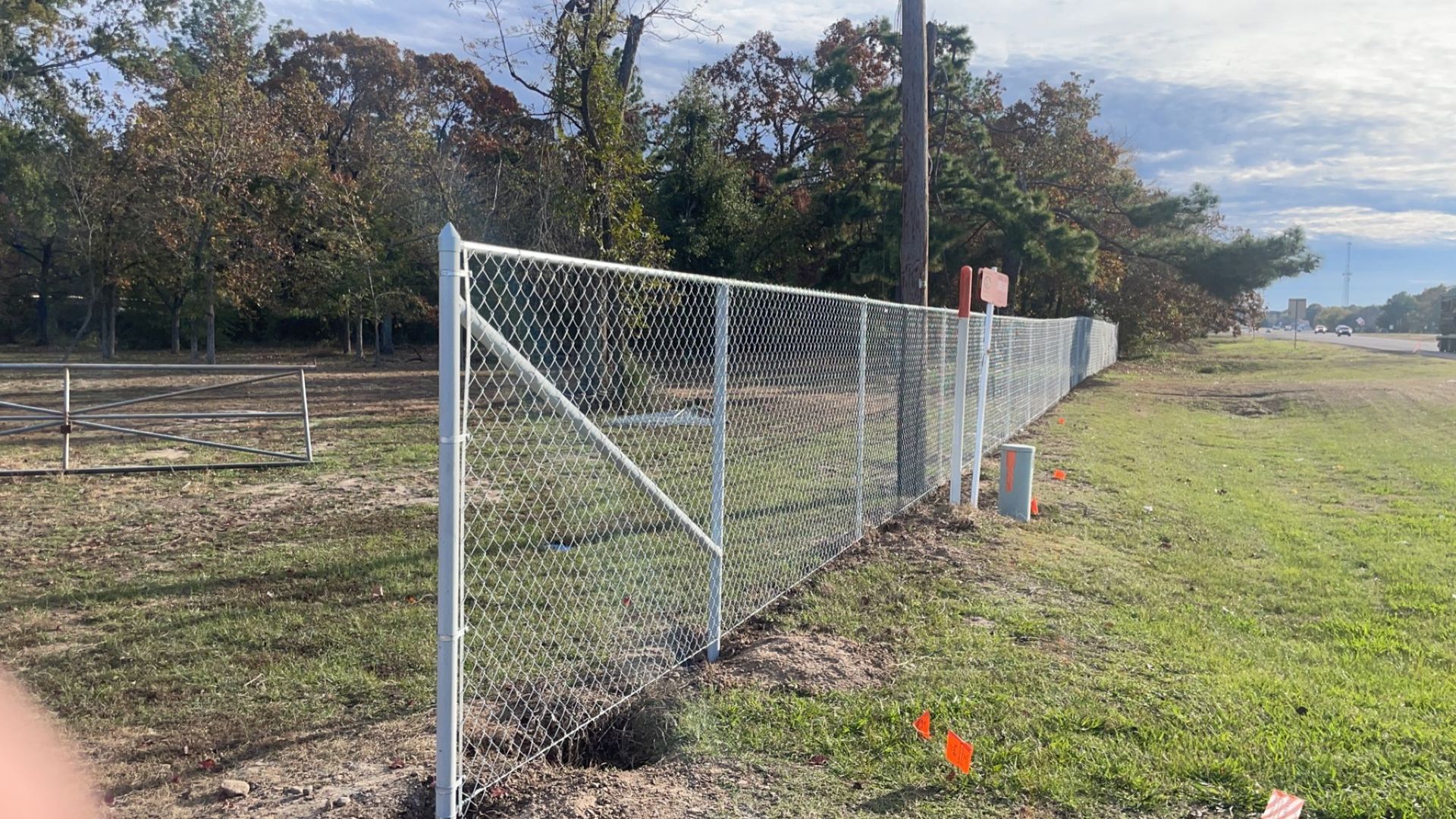 Chain-link fence along a grassy field beside trees under a cloudy sky.