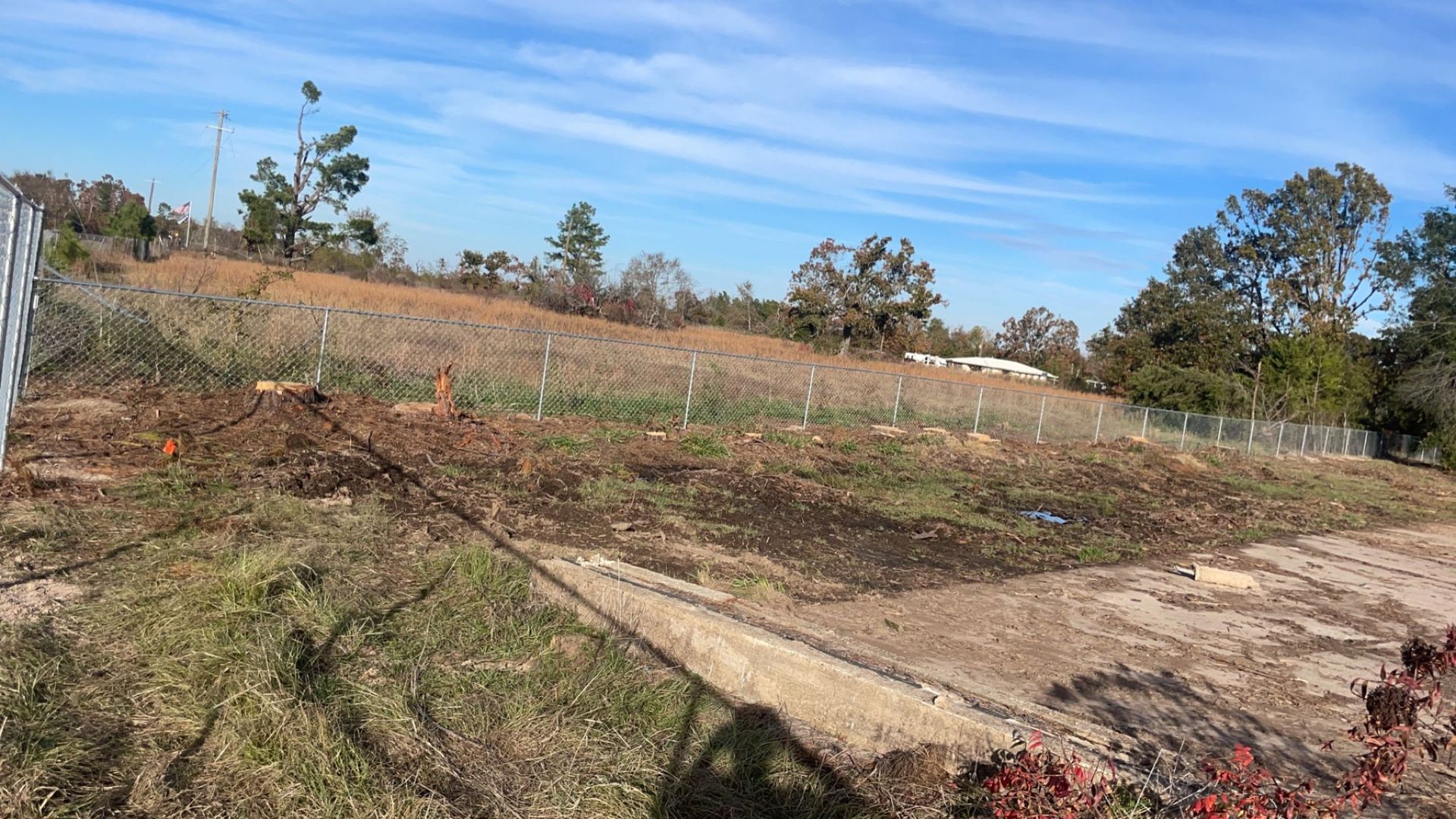 Dry, cleared field with sparse vegetation, dirt path, and trees under a blue sky