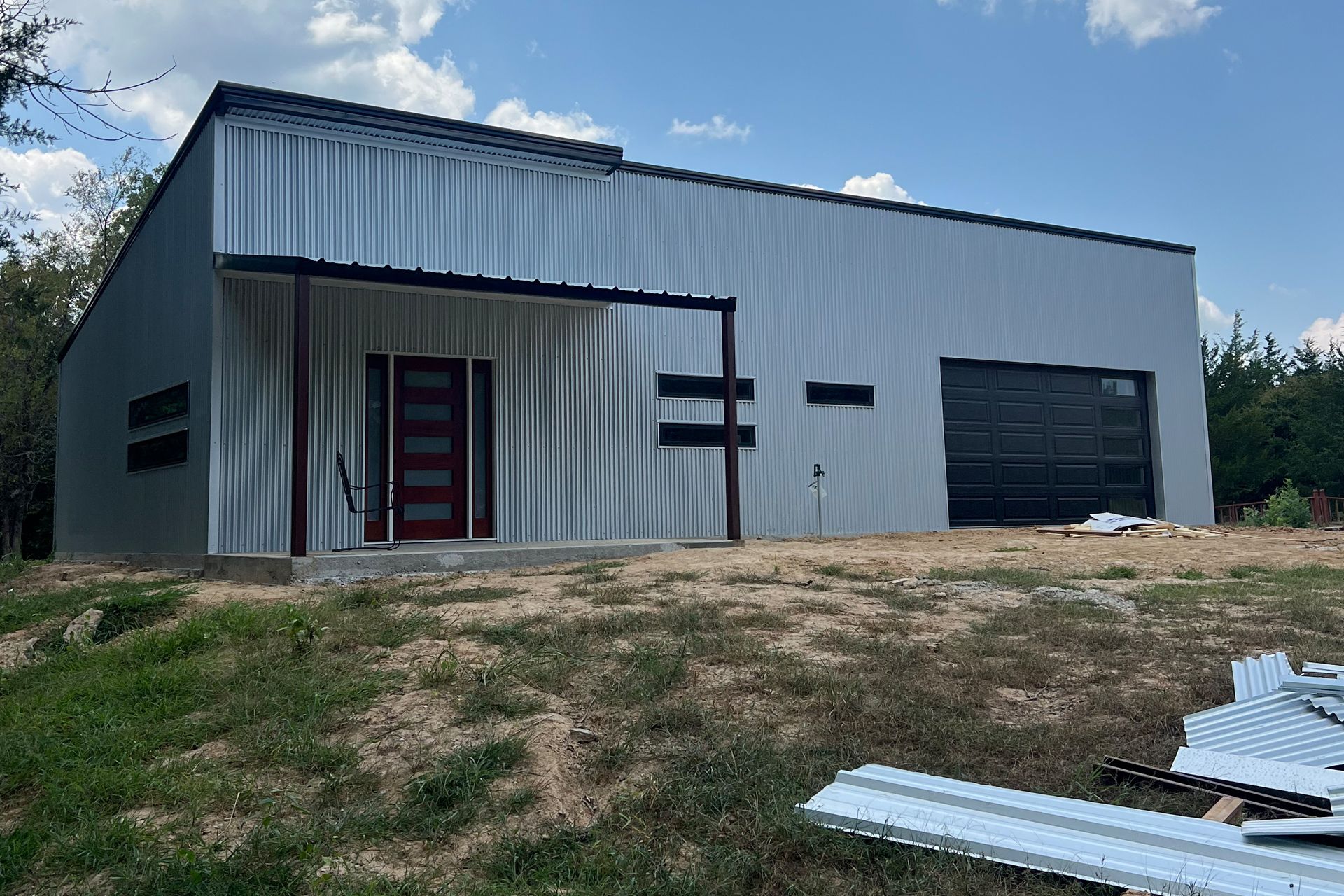 Gray metal warehouse building with red door, black garage door, and grassy foreground under blue sky