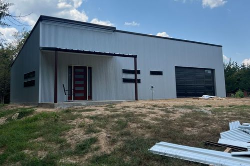 Gray metal warehouse building with red door, black garage door, and grassy foreground under blue sky
