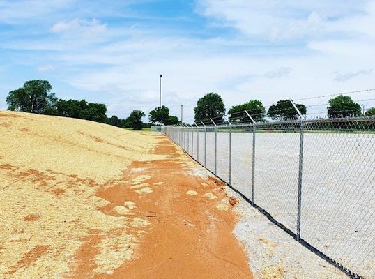 A chain link fence surrounds a dirt field with trees in the background.