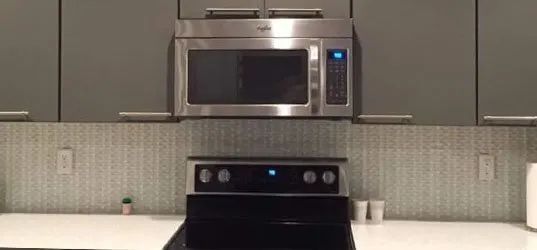 Kitchen with microwave over stove and gray cabinets. White tiled backsplash and countertop with a small plant.