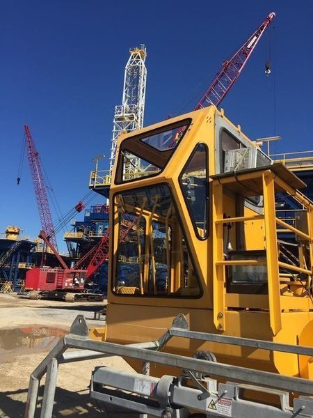 Yellow crane cab in the foreground at an oil rig with red and white cranes against a blue sky.