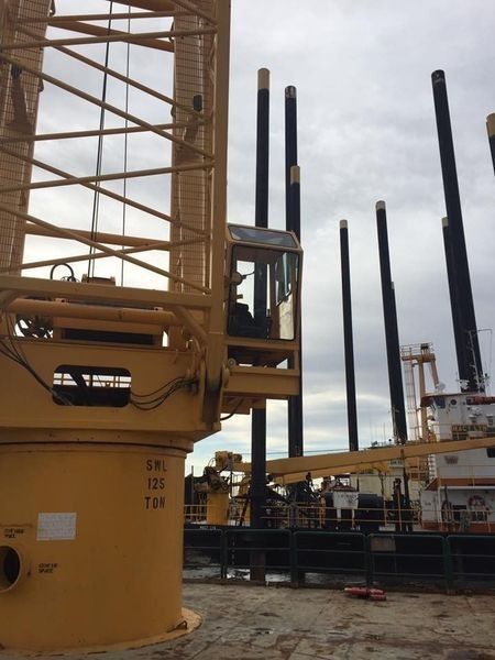 Yellow construction crane with a cab, operating near black pilings on a barge, cloudy sky.