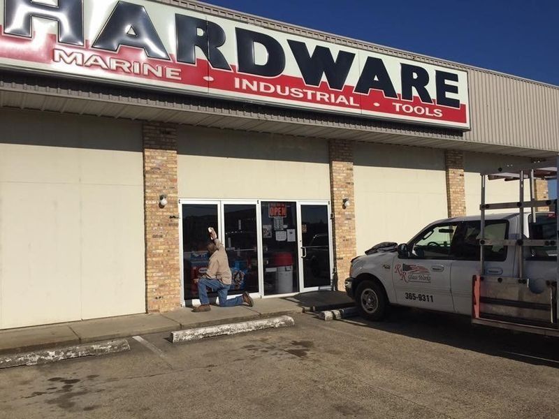 A man kneels in front of a hardware store entrance. A white truck is parked to the side.