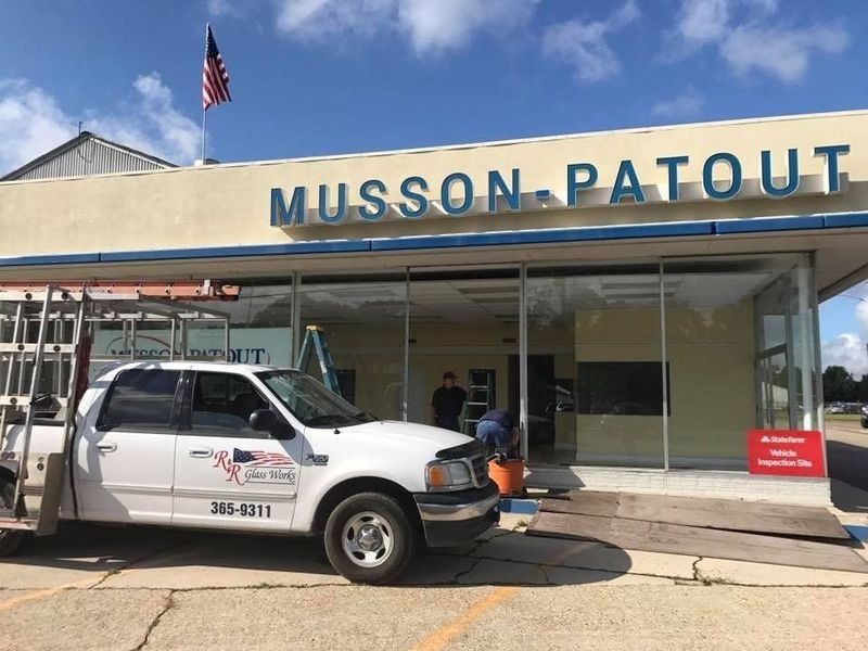 White truck at Musson-Patout building with workers installing glass. American flag flies.