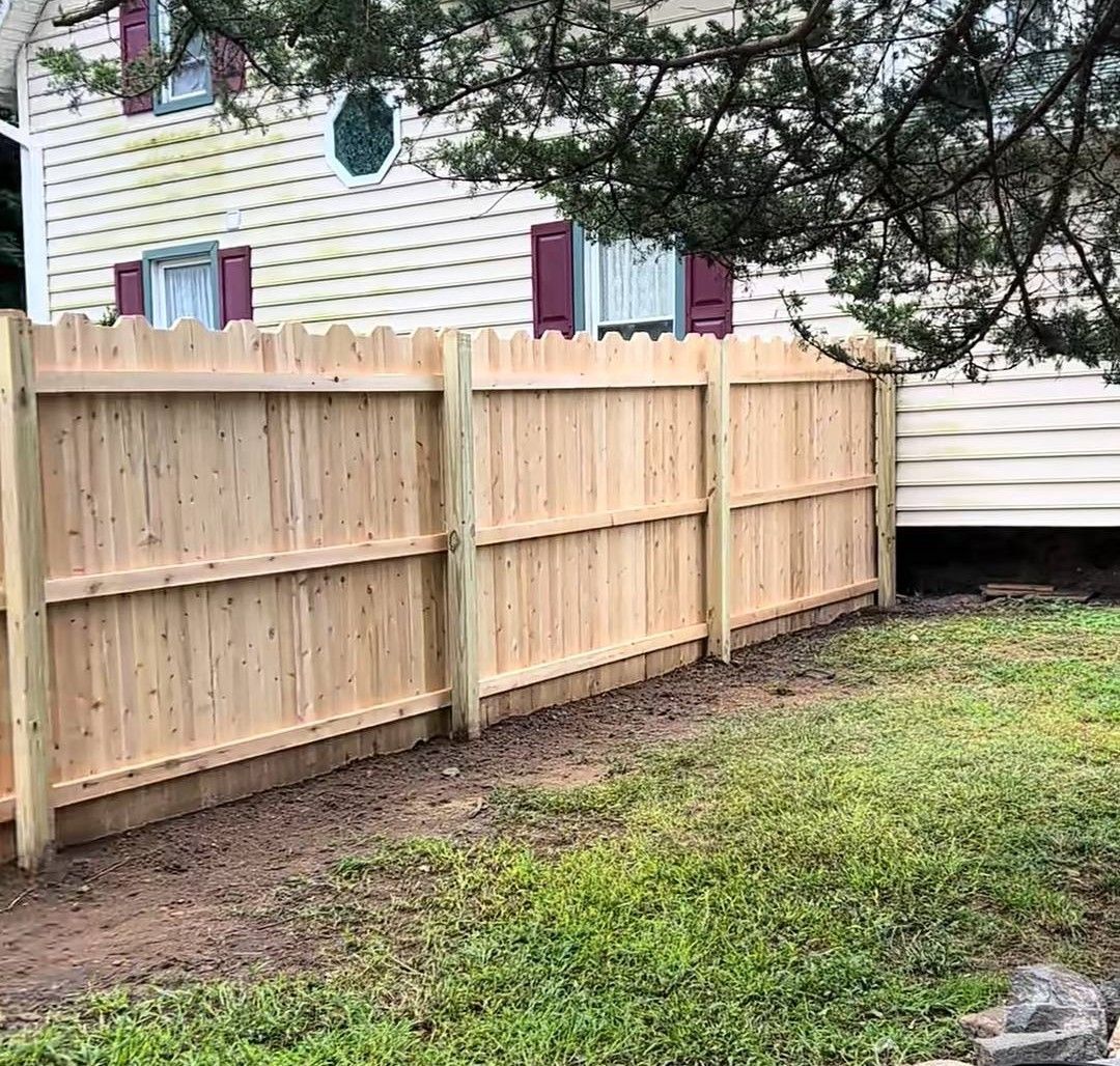 Wooden fence in front of a white house with burgundy shutters; the fence is in a yard with grass.