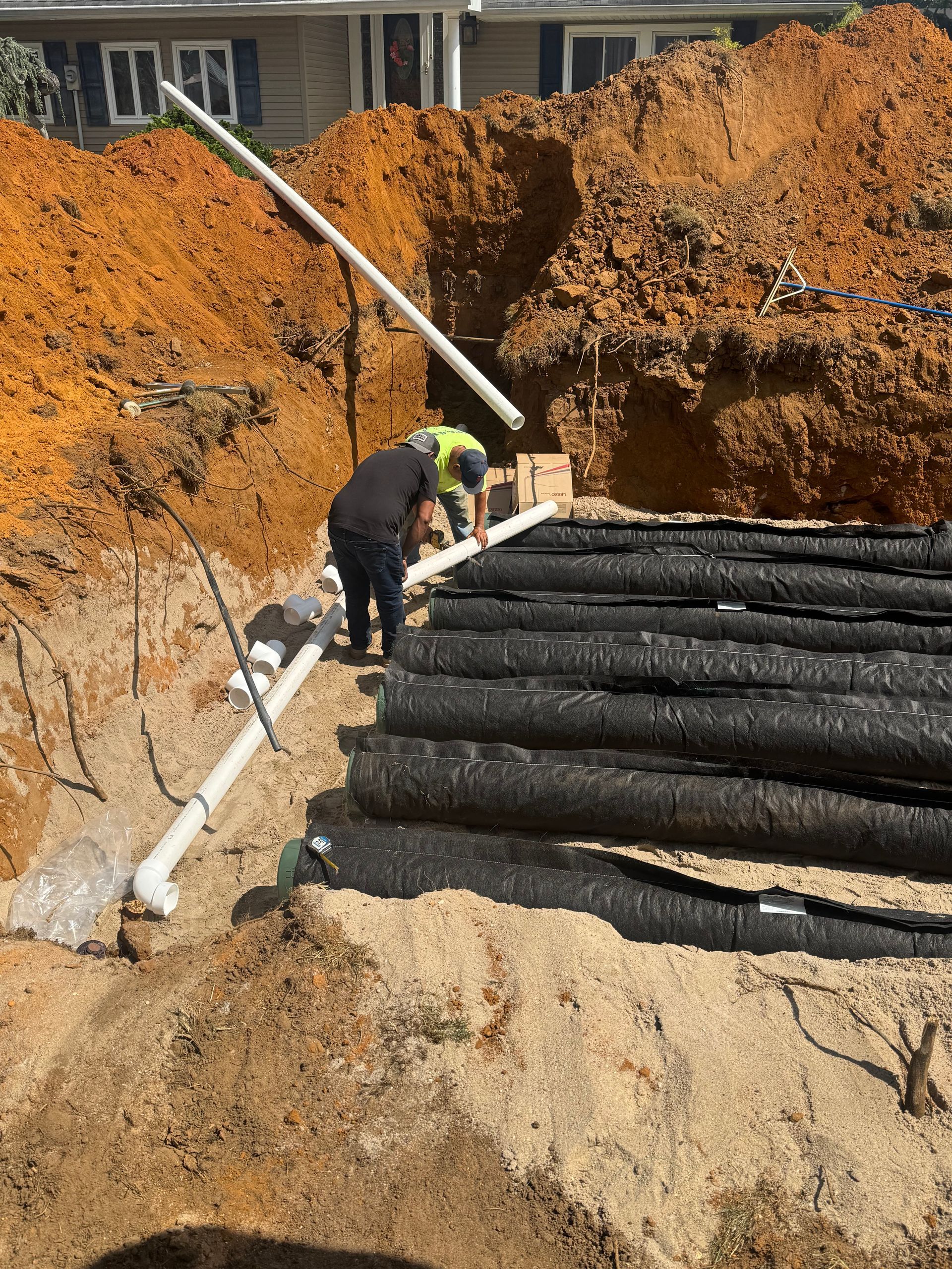 Workers installing drainage pipes in an excavated area with stacked, black tubes.