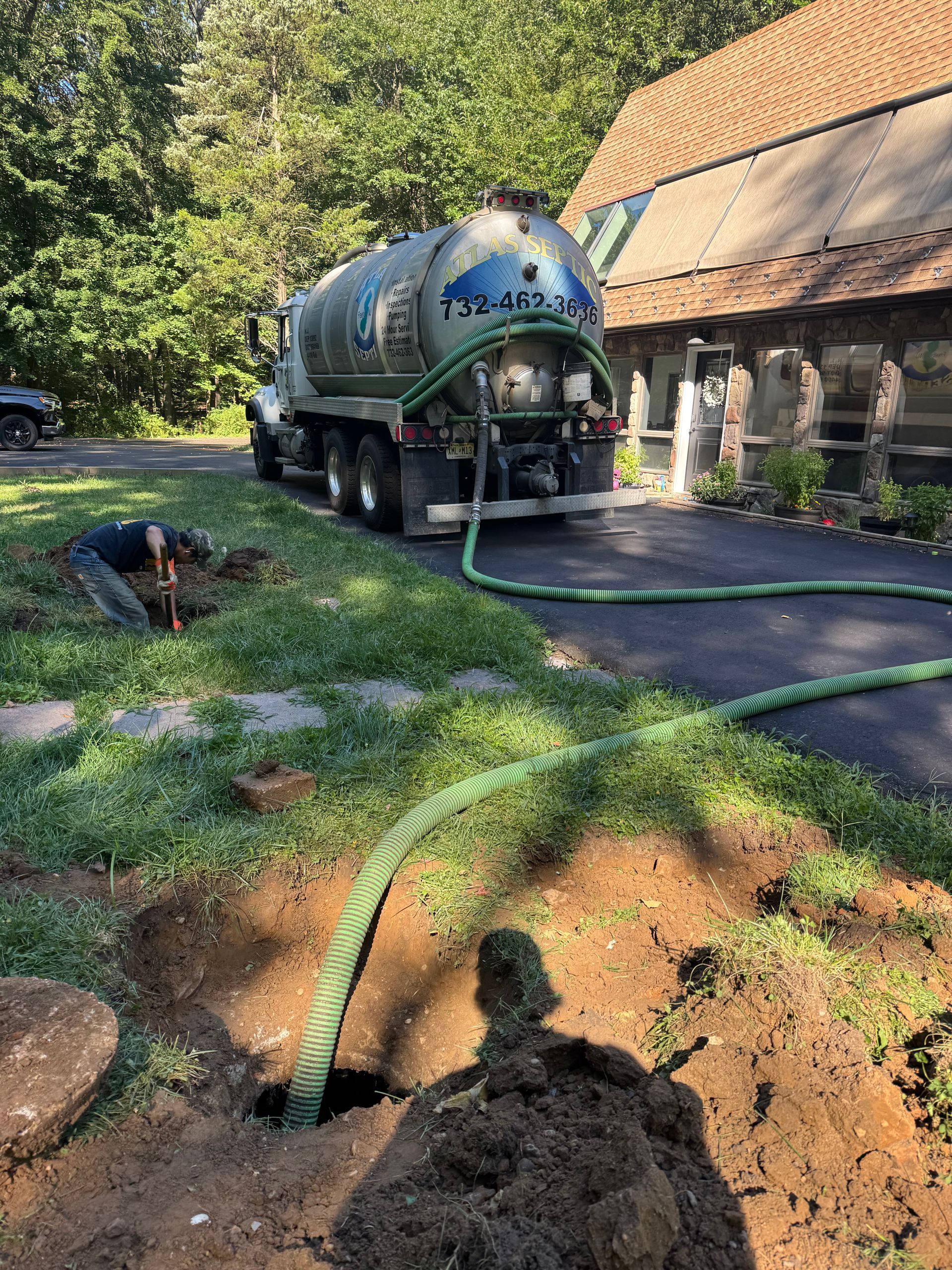 Septic tank pumping: Truck with hose connected to an open ground pit. A person is digging. House in the background.